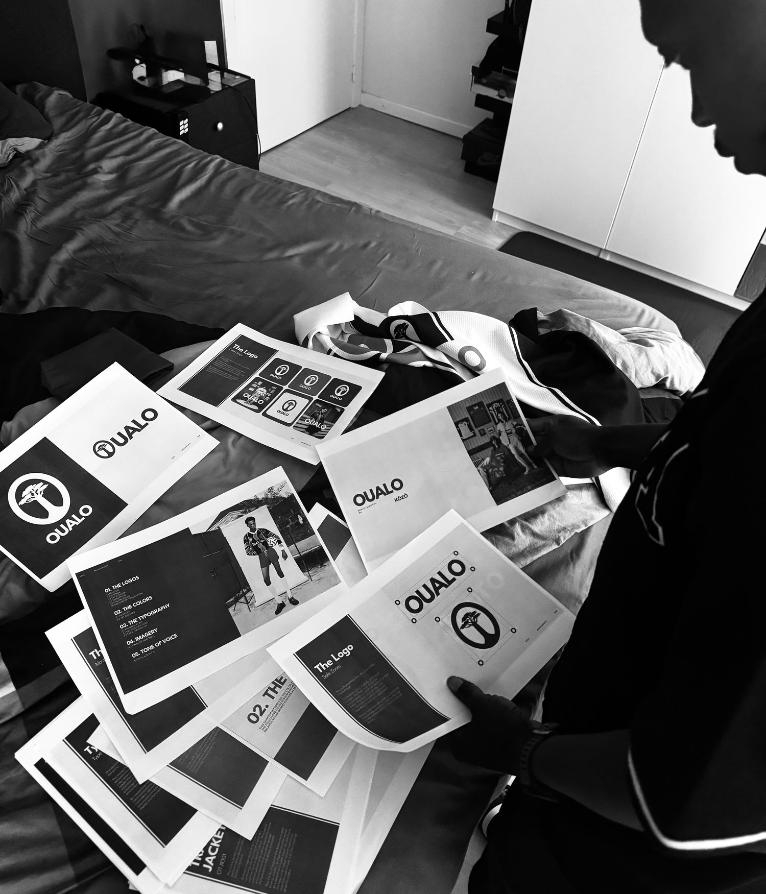 A person in a dark room looks at a pile of printed promotional materials and logos branded 'OUALO' spread across a bed.