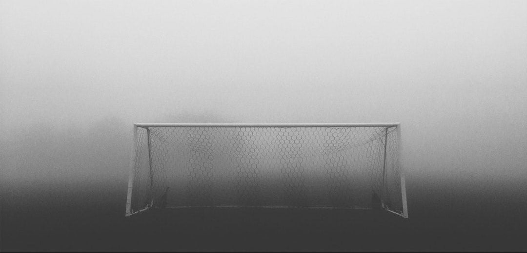 A soccer goal framed in white metal with a net, standing in heavy fog on a dark field. The scene is monochrome, with the goal appearing light gray against a misty, foggy background.