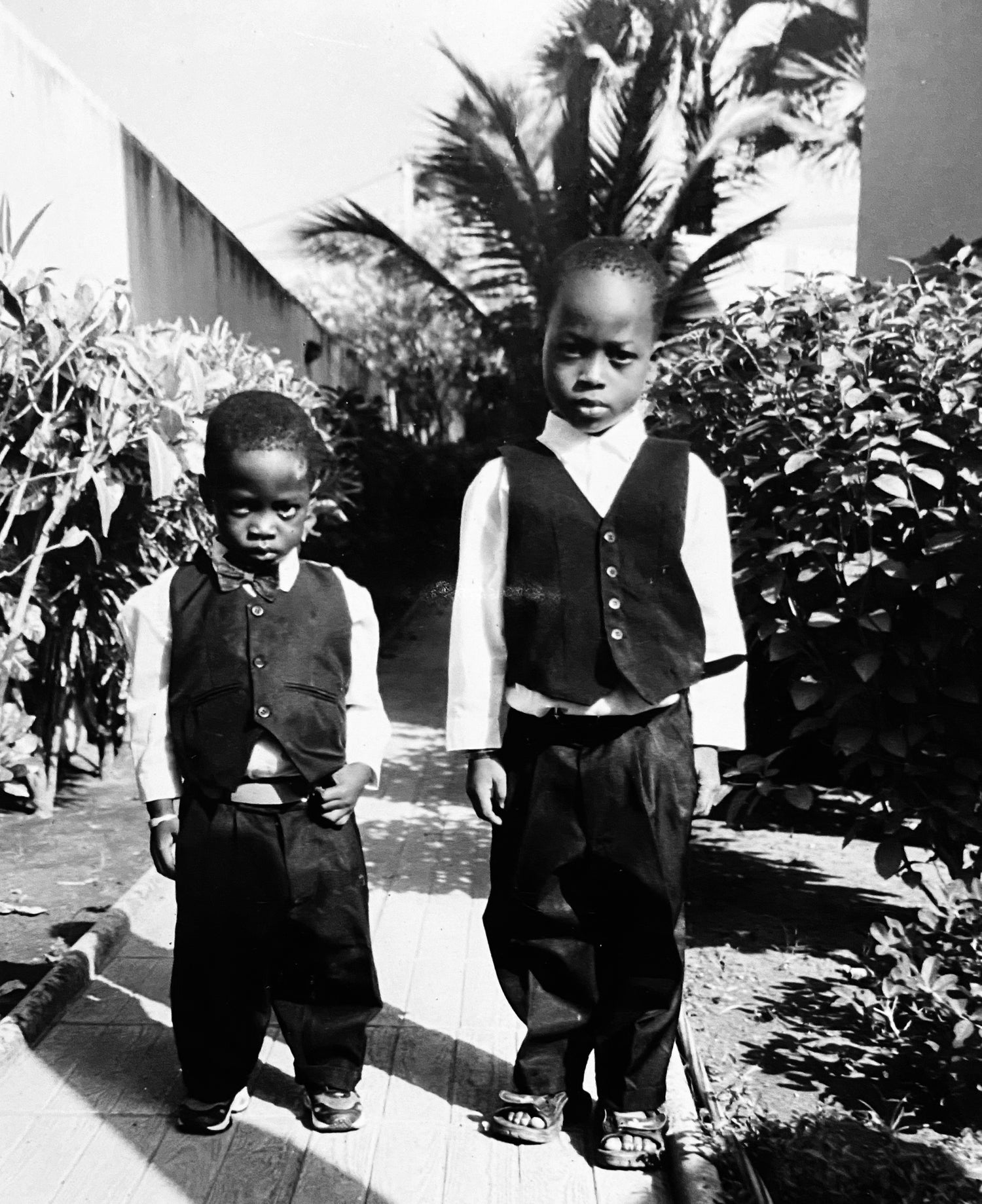 Black and white photograph of two young boys standing outdoors on a wooden path, wearing matching dark vests and trousers with light shirts.